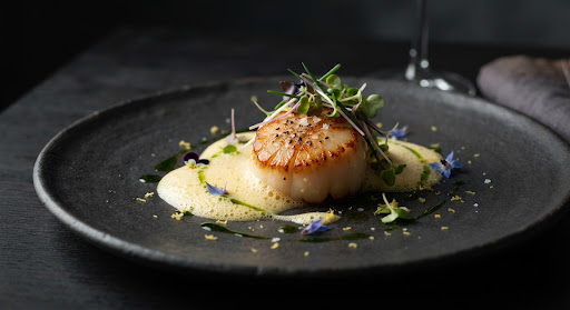 Beautifully plated Hokkaido scallop with saffron foam and microgreens on a dark ceramic plate.