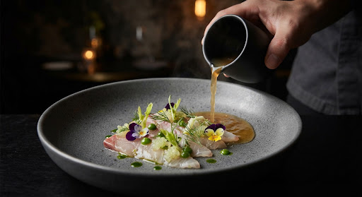 Seasonal tasting menu dish with cured fish, micro-herbs and an amber broth being poured.