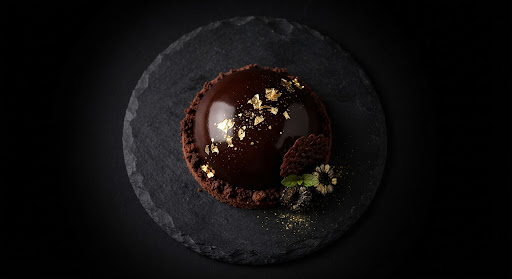 Dark chocolate dome dessert decorated with edible gold leaf on a textured stone plate.