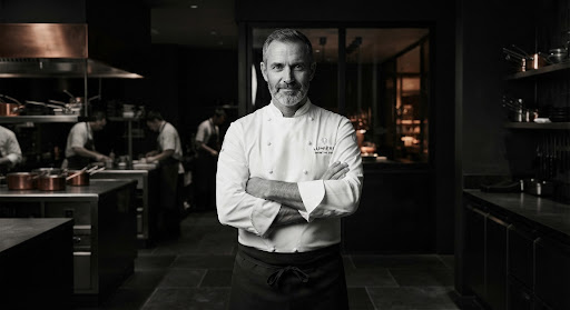 Black-and-white portrait of the executive chef in a white chef's coat in a minimalist kitchen.